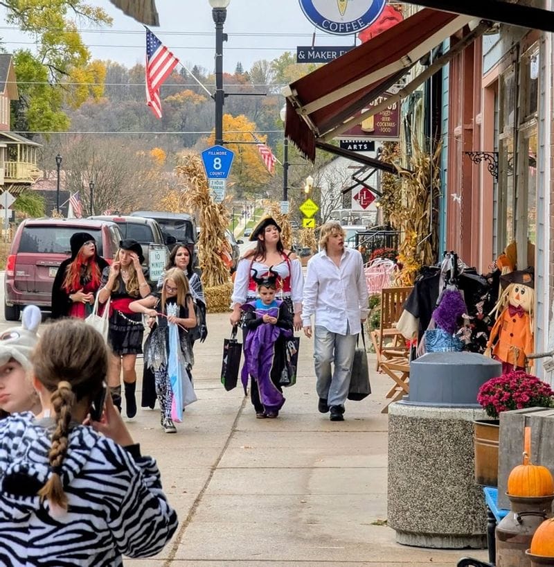 Families and children in Halloween costumes walking along a decorated downtown Lanesboro sidewalk during a business trick-or-treat event, with storefronts adorned in fall décor and pumpkins.