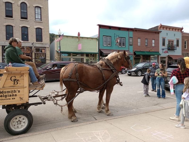 Horse-drawn wagon ride traveling through downtown Lanesboro during a fall event, with colorful historic storefronts lining the street and families gathered along the sidewalk.