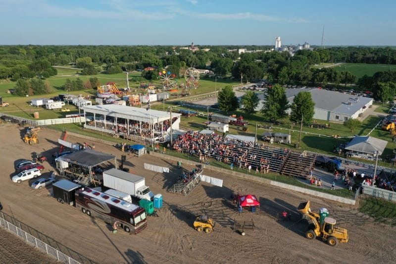 Aerial view of the Fillmore County Fairgrounds during a summer event, with grandstands filled with spectators, carnival rides in the background, and a concert stage set up along the dirt arena.