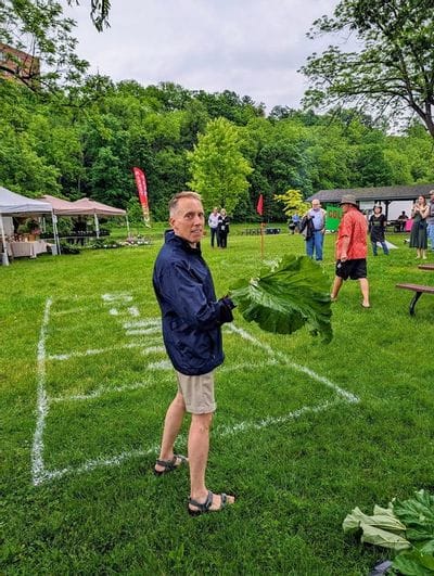 Man standing on a grassy field holding an oversized rhubarb leaf during an outdoor festival in Lanesboro, with tents, spectators, and wooded bluffs in the background.