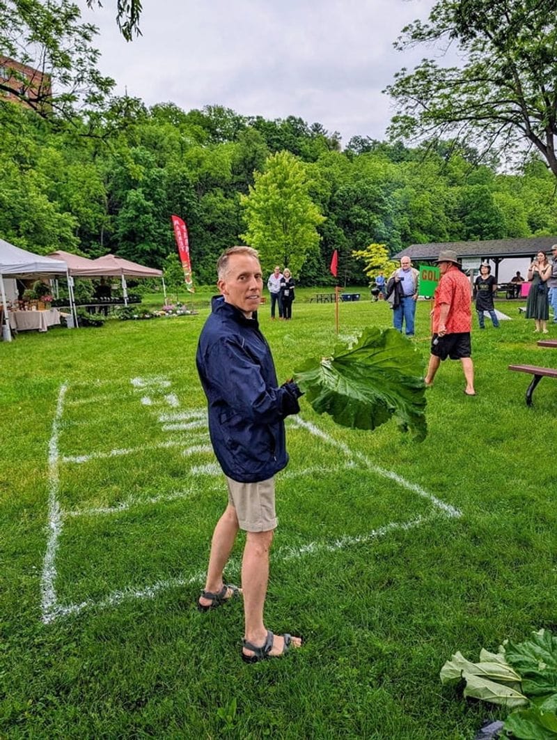 Man standing on a grassy field holding an oversized rhubarb leaf during an outdoor festival in Lanesboro, with tents, spectators, and wooded bluffs in the background.