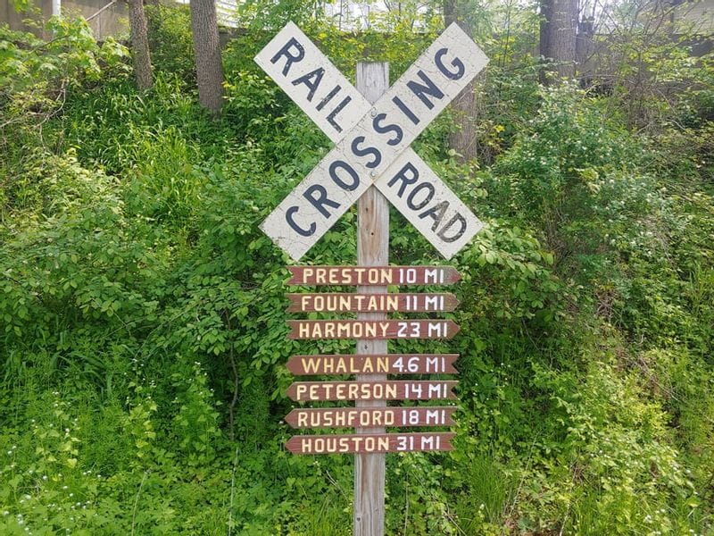 Railroad crossing sign along the Root River Trail with directional markers listing distances to Preston, Fountain, Harmony, Whalan, Peterson, Rushford, and Houston, surrounded by lush green vegetation.