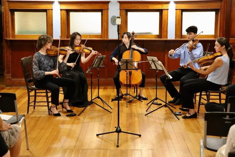 String quartet performing in an intimate indoor venue in Lanesboro, with two violinists, a violist, and a cellist seated on a wooden floor stage and audience members watching from nearby chairs.