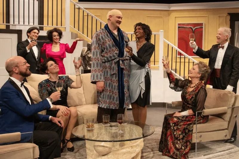 Actors perform a lively scene on stage at the Commonweal Theatre Company in Lanesboro, raising glasses in an elegant living-room setting during a theatrical production, with performers dressed in formal and vintage-inspired costumes.
