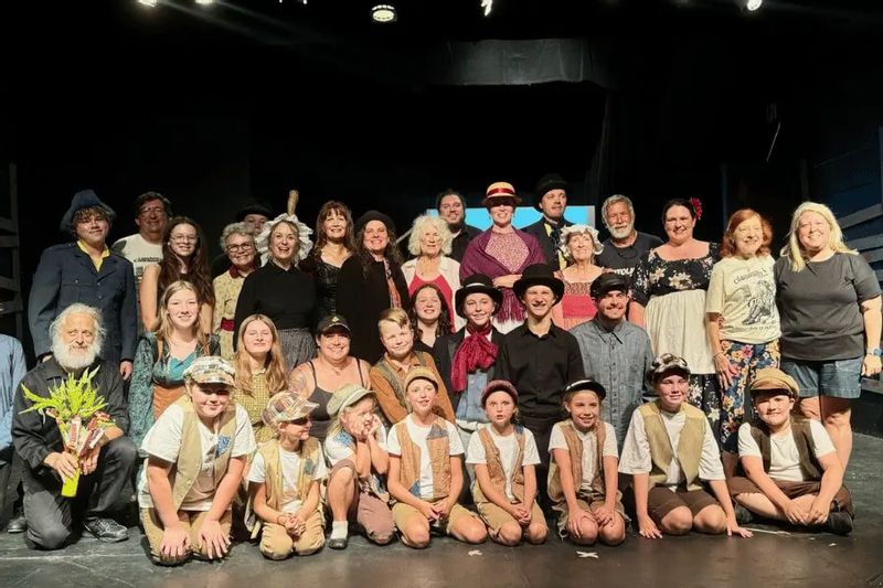 Cast members of a community theater production pose together on stage after a performance in Lanesboro, featuring adults and children in period-style costumes, celebrating a successful show under stage lights.