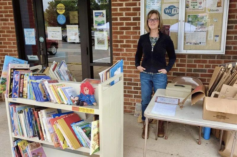 A volunteer stands outside the Lanesboro Public Library next to a book sale organized by Friends of the Lanesboro Library, with shelves of children’s books and tables of donated items set up on the sidewalk.