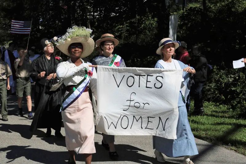 Participants in period clothing march during a History Alive! living history event in Lanesboro, carrying a banner that reads “Votes for Women” as part of a women’s suffrage reenactment.