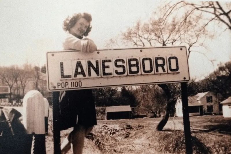 A historic photograph shows a young woman standing beside a roadside sign reading “Lanesboro, Pop. 1100,” capturing an early glimpse of the town’s past with homes and trees in the background.