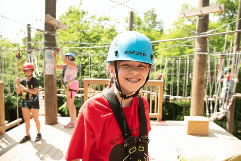A smiling camper wearing a helmet and safety harness stands on a high ropes course at Eagle Bluff Environmental Learning Center, with other participants navigating rope obstacles among the trees in the background.
