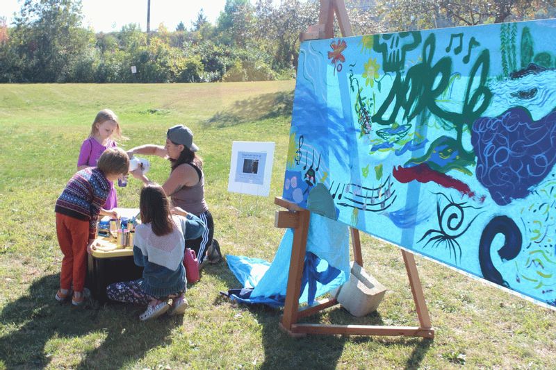 Children and an instructor create a collaborative outdoor painting during a community art activity hosted by Lanesboro Arts, with a colorful river-themed mural displayed on an easel in a grassy park setting.