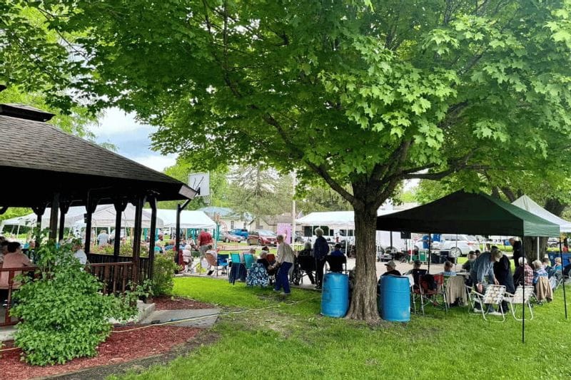 People gather under shade trees and canopy tents near a park gazebo in Whalan, Minnesota, enjoying live music and community activities on a summer day.