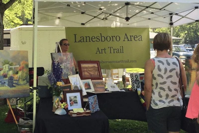 An artist booth at the Lanesboro Area Art Trail features paintings, framed artwork, and handmade pieces displayed under a white tent as visitors browse during a summer arts event.