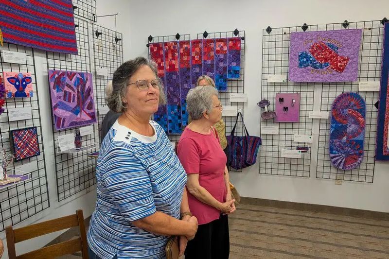 Visitors stand inside the Driftless Fiber Arts Collective gallery in Lanesboro, viewing colorful textile and fiber art pieces displayed on wall grids.