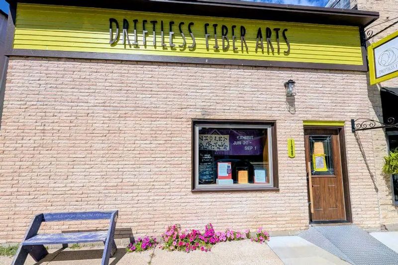 Exterior of the Driftless Fiber Arts Collective in downtown Lanesboro, featuring a yellow sign above a brick storefront with display window, wooden door, and flowers along the sidewalk.