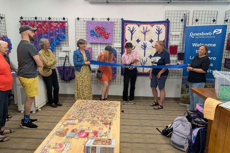 Community members and organizers celebrate a ribbon cutting at the Driftless Fiber Arts Collective in Lanesboro, standing beneath colorful quilt and fiber art displays with a community puzzle table in the foreground.