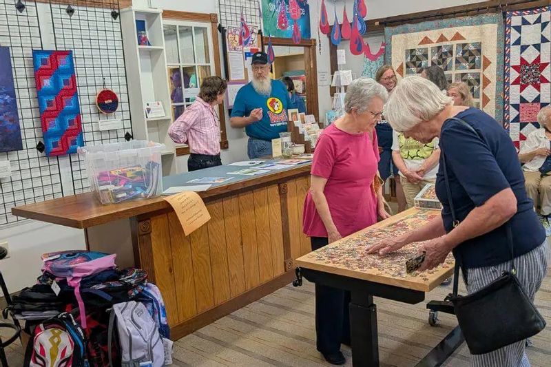 Visitors gather inside the Driftless Fiber Arts Collective in Lanesboro, working on a community puzzle while surrounded by colorful quilts and fiber art displays.