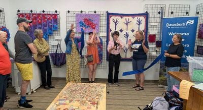 Community members celebrate a ribbon cutting at the Driftless Fiber Arts Collective in Lanesboro, with quilts displayed on the wall and attendees clapping during the opening ceremony.