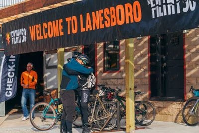 Two cyclists hug beneath a “Welcome to Lanesboro” Filthy 50 banner outside Sylvan Brewing, with gravel bikes leaned nearby after the race.