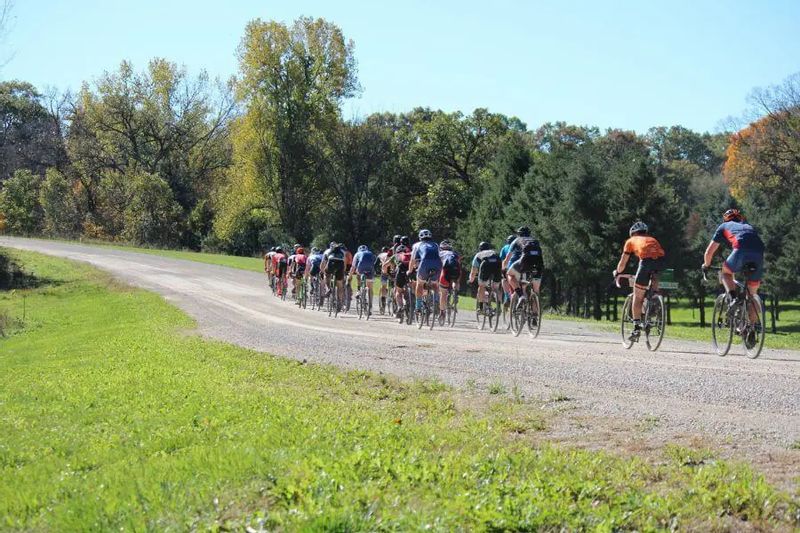 A group of cyclists rides together along a scenic gravel road during the Filthy 50, surrounded by green fields and trees just beginning to show fall color in the Lanesboro countryside.