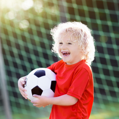 Young child with curly blonde hair smiling and holding a soccer ball while standing in front of a soccer goal on a sunny day.