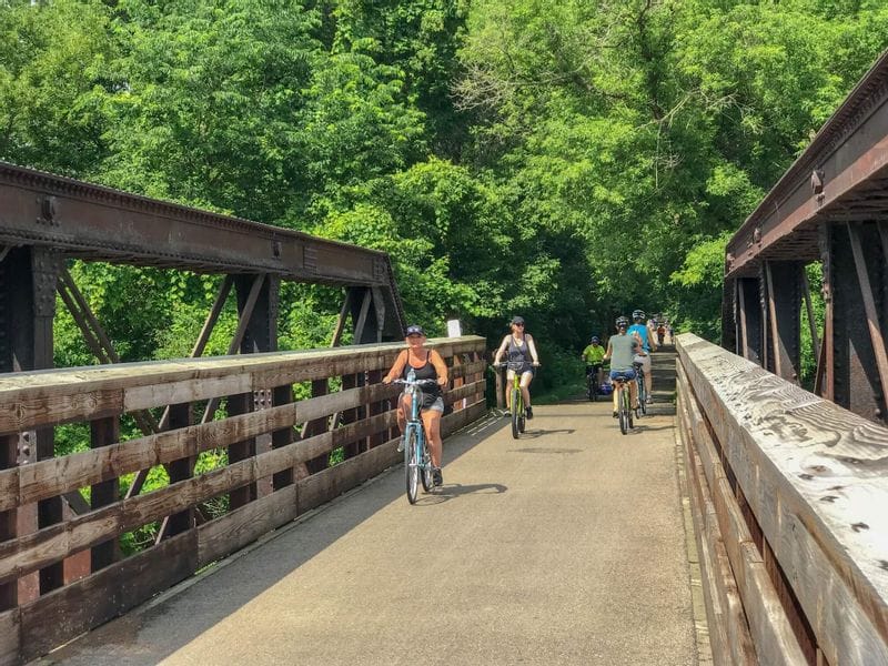 Cyclists ride across a historic steel bridge on the Root River Trail in Lanesboro, Minnesota, surrounded by lush green trees on a summer day.