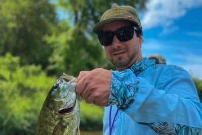 Angler wearing sunglasses and a blue jacket holds a freshly caught smallmouth bass along a green, tree-lined river on a sunny day.