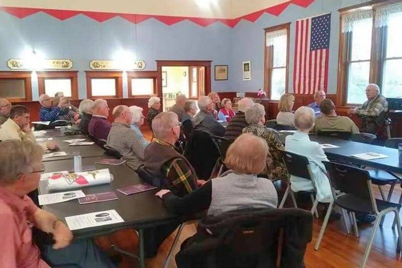 Community members sit at tables inside a historic lodge hall in Lanesboro, listening to a speaker during a Sons of Norway gathering, with an American flag hanging on the wall.