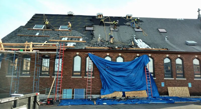 Construction crews work on a steep roof of a large brick building, removing and replacing shingles while secured with safety ropes, with ladders, scaffolding, and blue tarps covering part of the structure below.