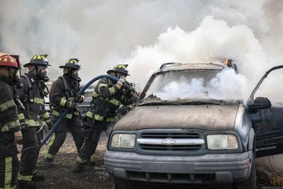 Several firefighters in full protective gear and breathing masks work together to put out a vehicle fire. One firefighter sprays water into the front of a heavily smoking SUV while others assist nearby with the hose line. Thick white smoke rises from the vehicle, and small flames are visible near the roof. The scene appears to be part of a fire response or live training exercise.