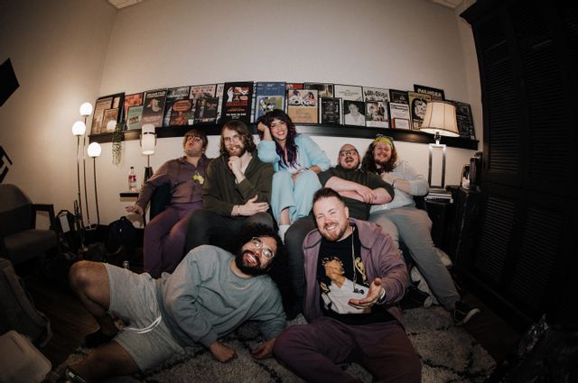 The band Florencia and the Feeling gathers together in a relaxed backstage-style room, posing casually for a group photo. Some members sit on a couch while others sit on the floor, smiling and interacting with the camera. Music posters line the wall behind them, creating a creative and welcoming atmosphere connected to live music and performance.