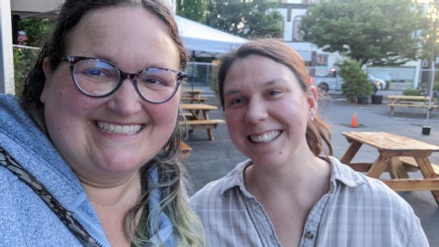 Two Caucasian women looking at the camera from close-up. Both with brown hair and smiling. There are picnic tables and a white canopy in the background.