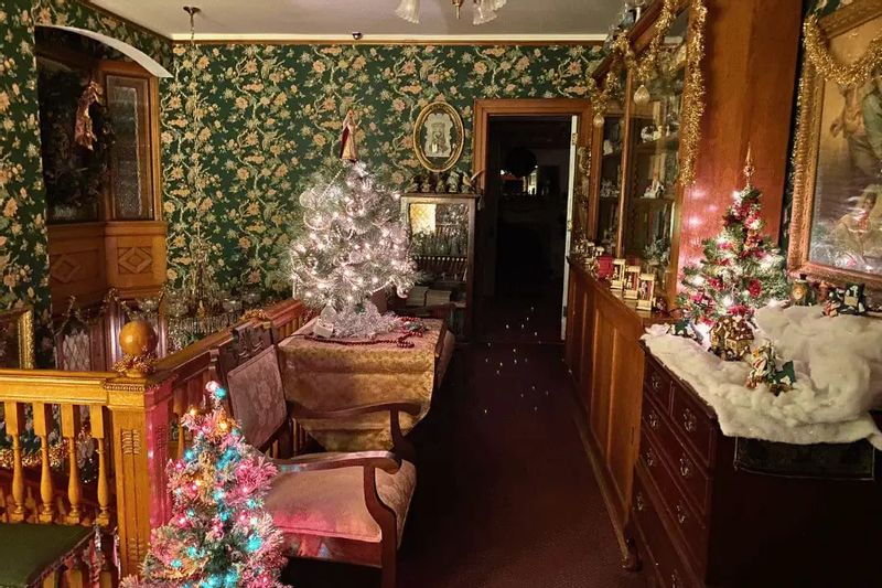 Victorian-style interior at Oakenwald Terrace decorated for the holidays, featuring floral wallpaper, antique furniture, and multiple small Christmas trees glowing with vintage lights.