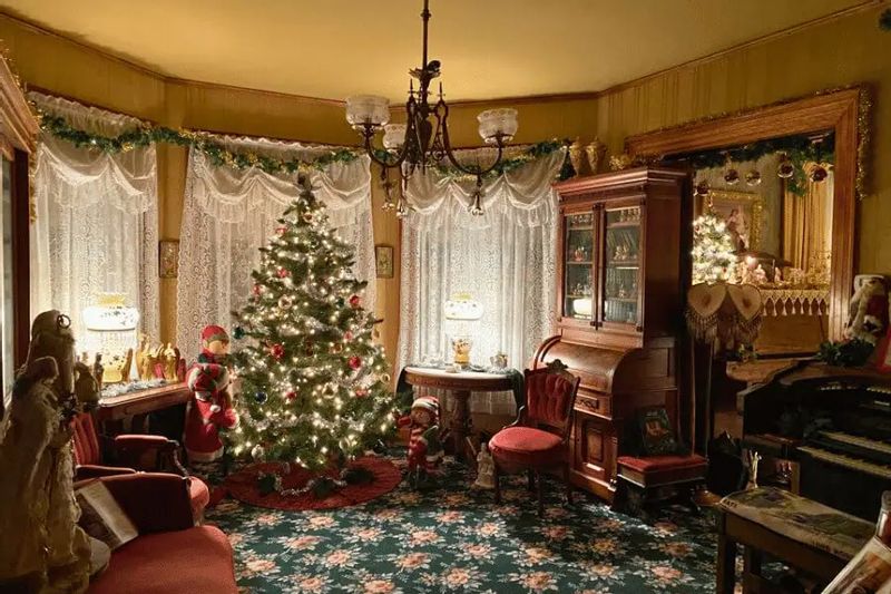 A Victorian-style parlor at Oakenwald Terrace in Chatfield decorated for the holidays, featuring a lit Christmas tree, antique furniture, lace curtains, and festive garlands in a warm, historic setting.