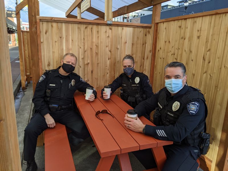 Three Hillsboro Police Officers in uniform sitting at an orange picnic table in a wooden fenced parklet in front of Insomnia Coffee Company. 