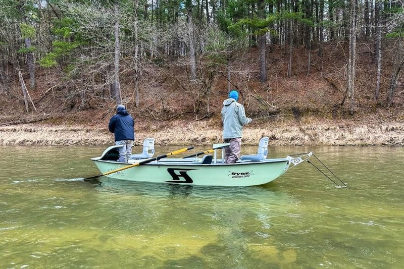 Two anglers fish from a drift boat on a calm river during a guided trip with Root River Rod Co., with a wooded shoreline in the background.