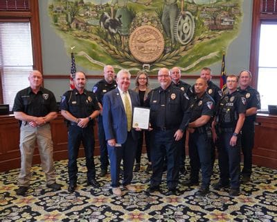 Statesville Police Chief Onley, members of the Statesville Police Department, and Mayor Doug Hendrix pose together inside City Hall following the presentation of a proclamation recognizing May 11–16, 2026 as National Police Week in the City of Statesville. Chief Onley stands beside Mayor Hendrix while holding the framed proclamation, surrounded by uniformed officers and department staff beneath the City seal mural in the council chambers.