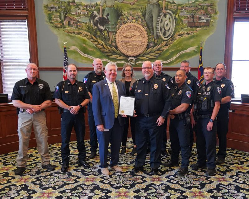 Statesville Police Chief Onley, members of the Statesville Police Department, and Mayor Doug Hendrix pose together inside City Hall following the presentation of a proclamation recognizing May 11–16, 2026 as National Police Week in the City of Statesville. Chief Onley stands beside Mayor Hendrix while holding the framed proclamation, surrounded by uniformed officers and department staff beneath the City seal mural in the council chambers.