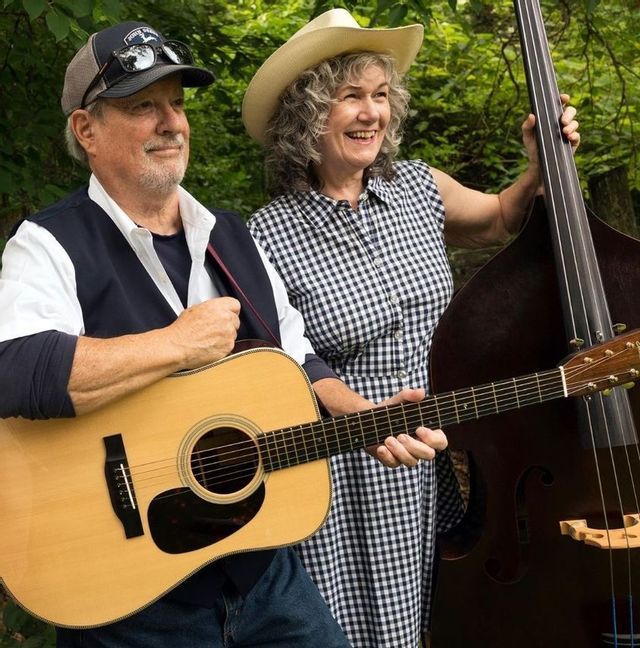 A man is holding a guitar and a woman is holding another stringed insturment. They are in front of a bush and are looking off to the right.