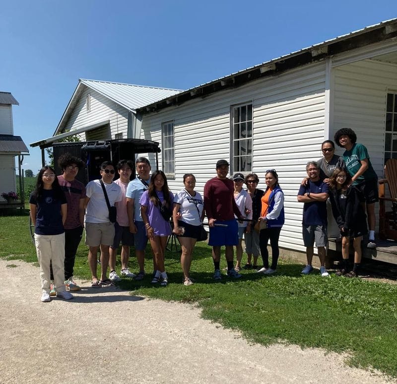 Group of visitors standing outside a white farm building during a Bluffscape Amish tour, gathered on a gravel driveway beside green grass under a clear blue sky, with rural farmland and outbuildings in the background.