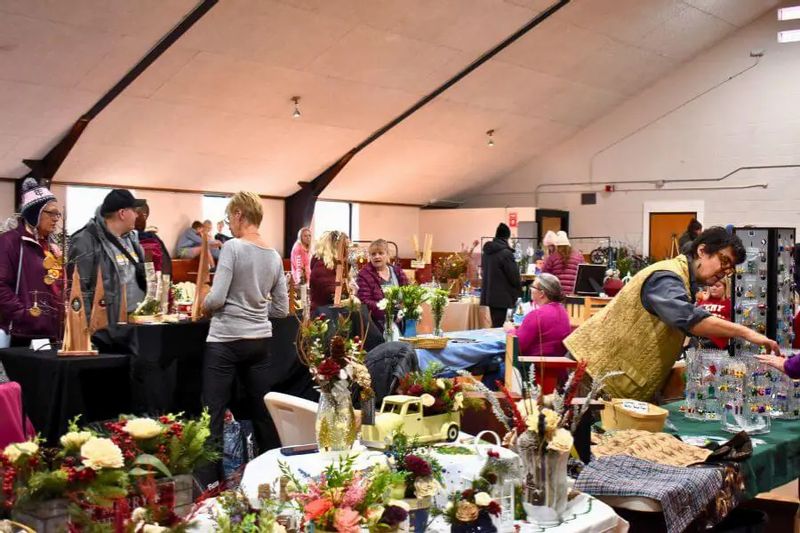 Visitors browse vendor tables filled with handmade goods, floral arrangements, and crafts inside a busy indoor winter market, with people chatting and shopping throughout the room.