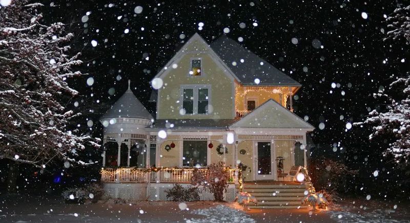 Snow falls at night around a warmly lit Scandinavian Inn in Lanesboro, with holiday lights glowing on the porch and roof, creating a cozy winter scene against the dark sky.
