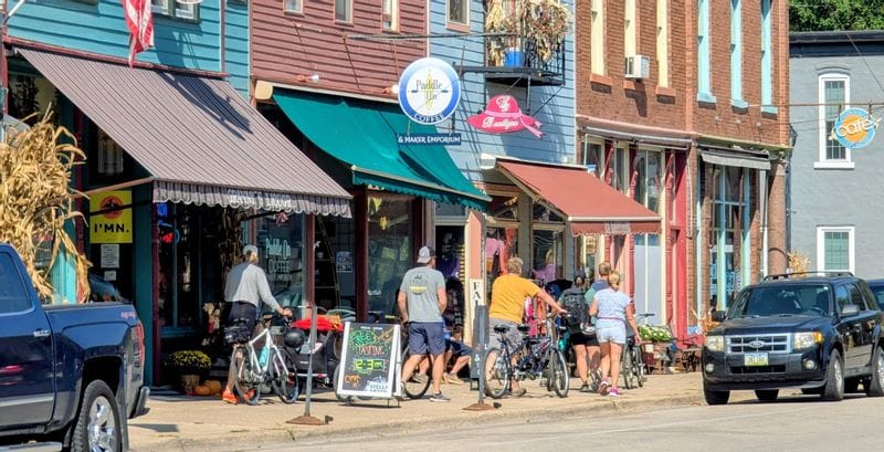 Shoppers and cyclists browse colorful downtown storefronts in Lanesboro, Minnesota, with local shops, cafés, and bikes lined along the sidewalk.