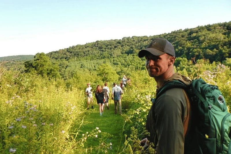 Hikers walk along a grassy trail through wildflowers at Eagle Bluff Environmental Learning Center near Lanesboro, with a guide carrying a backpack in the foreground on a sunny day.