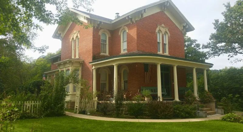 A historic red brick Victorian home with a wraparound porch and decorative details sits among mature trees and gardens, showcasing the Thompson House in Lanesboro, Minnesota.