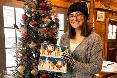 A smiling woman at Stone Mill Gifts holds a holiday-themed gift box beside a decorated Christmas tree inside the cozy shop during the holiday season.