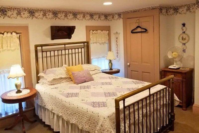 Cozy guest bedroom at the James A. Thompson House Bed & Breakfast, featuring a brass bed frame with a patterned quilt, vintage bedside tables with lamps, soft pastel trim, and antique furnishings in a historic Victorian-style room.