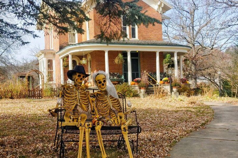 Two Halloween skeleton decorations dressed as a couple sit on a bench in front of the James A. Thompson House Bed & Breakfast, with the historic brick home, wraparound porch, and autumn foliage in the background.