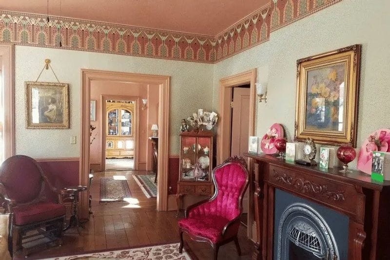 Victorian-style interior of the James A. Thompson House Bed & Breakfast, featuring ornate wallpaper borders, antique furniture, a decorative fireplace, framed artwork, and pink upholstered chairs in a historic parlor.