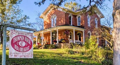 Exterior of the James A. Thompson House Bed & Breakfast in Lanesboro, Minnesota, a historic red-brick home with a wraparound porch, autumn decorations, and a sign in the front yard.
