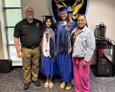 Police Chief Onley and Community Resource Coordinator Nakayla Griffin stand with SPD Junior Ambassadors Andrea Ayala and Tiffany Dubon inside the Statesville Police Department. Andrea and Tiffany are dressed in blue graduation caps and gowns with honor cords and medals as they celebrate their graduation achievements.